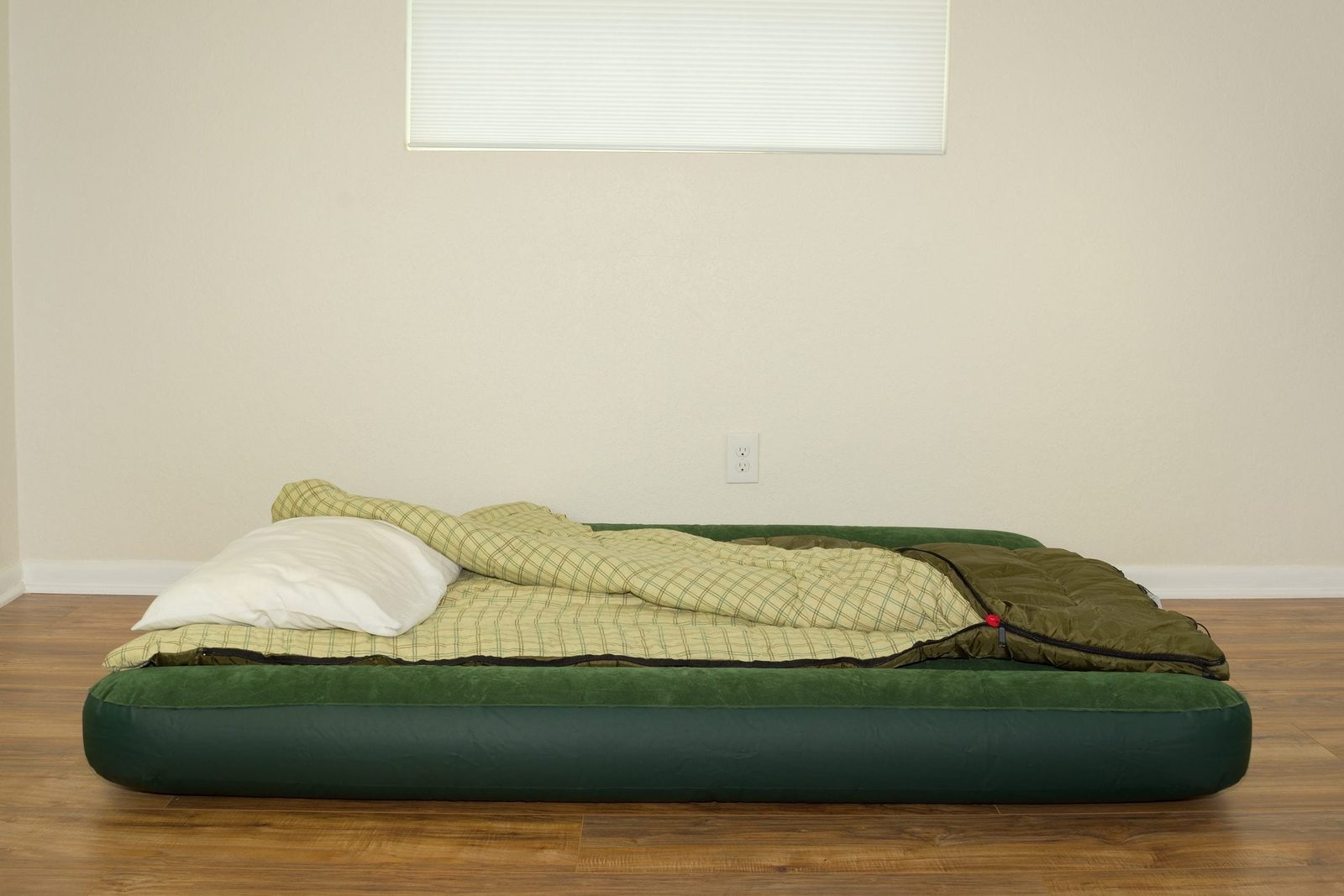 An inflatable mattress rests on wooden flooring, covered with a plaid blanket and a white pillow, in a minimally decorated room with a small