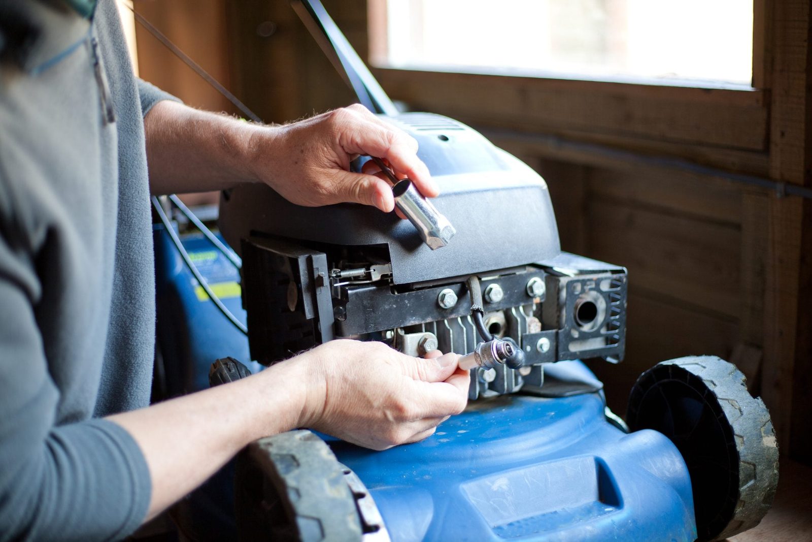 A person is replacing a spark plug on a blue lawn mower inside a wooden shed, using a socket wrench and focusing on the engine.