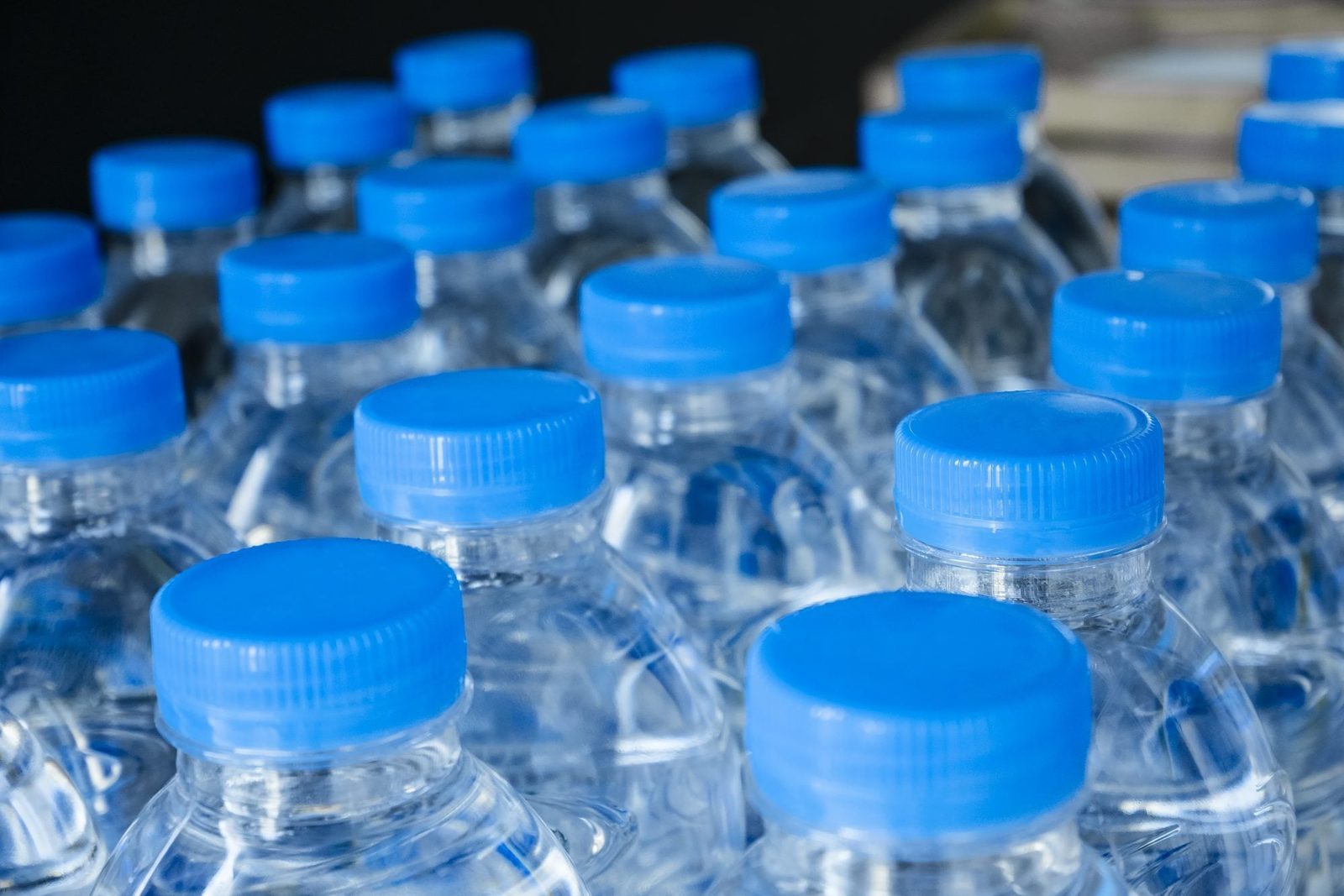 Clear plastic water bottles with blue caps are arranged closely together, creating a uniform pattern against a dark background.