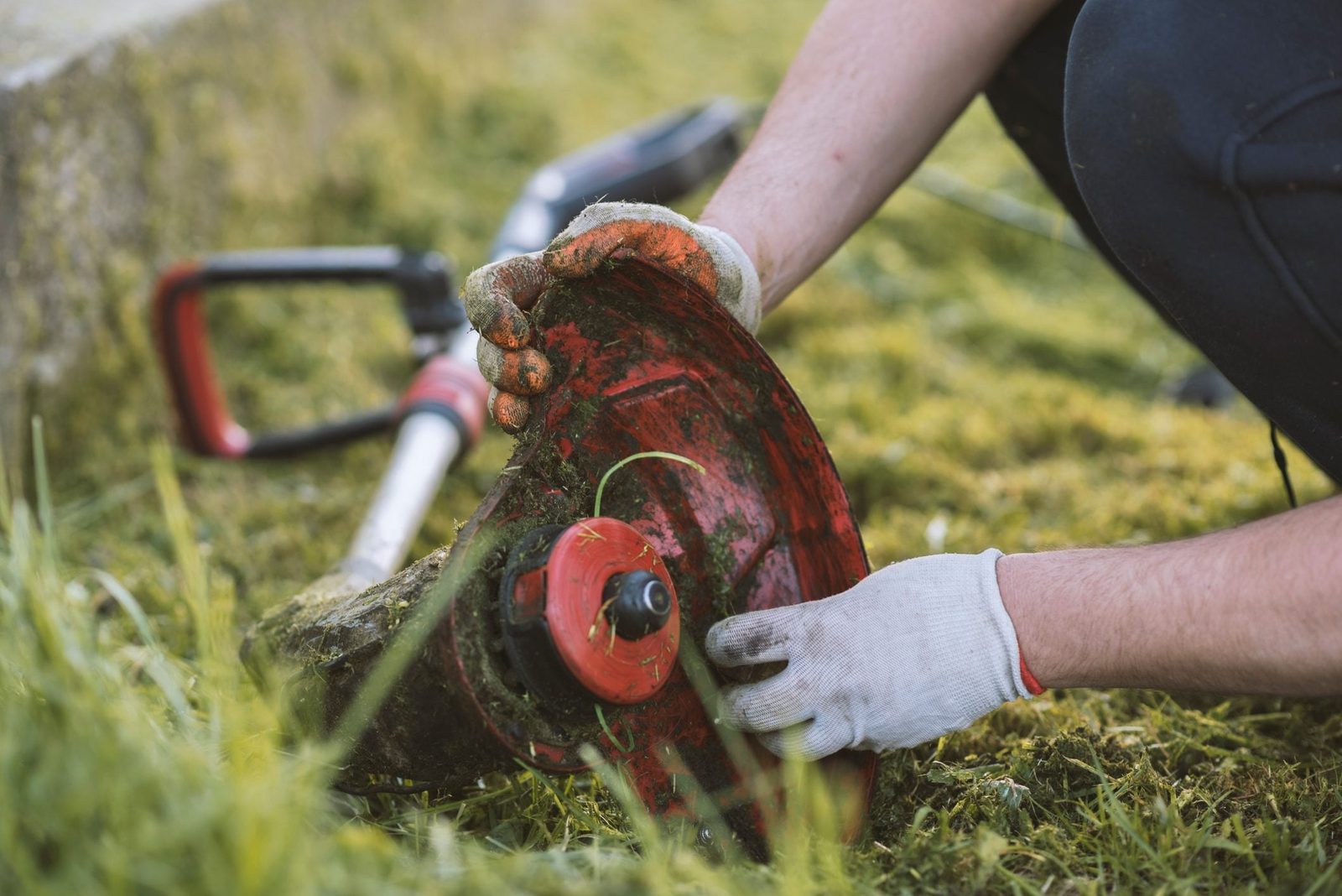 A person adjusts a red lawn equipment spool while kneeling in grass, surrounded by green foliage and tools for yard maintenance.