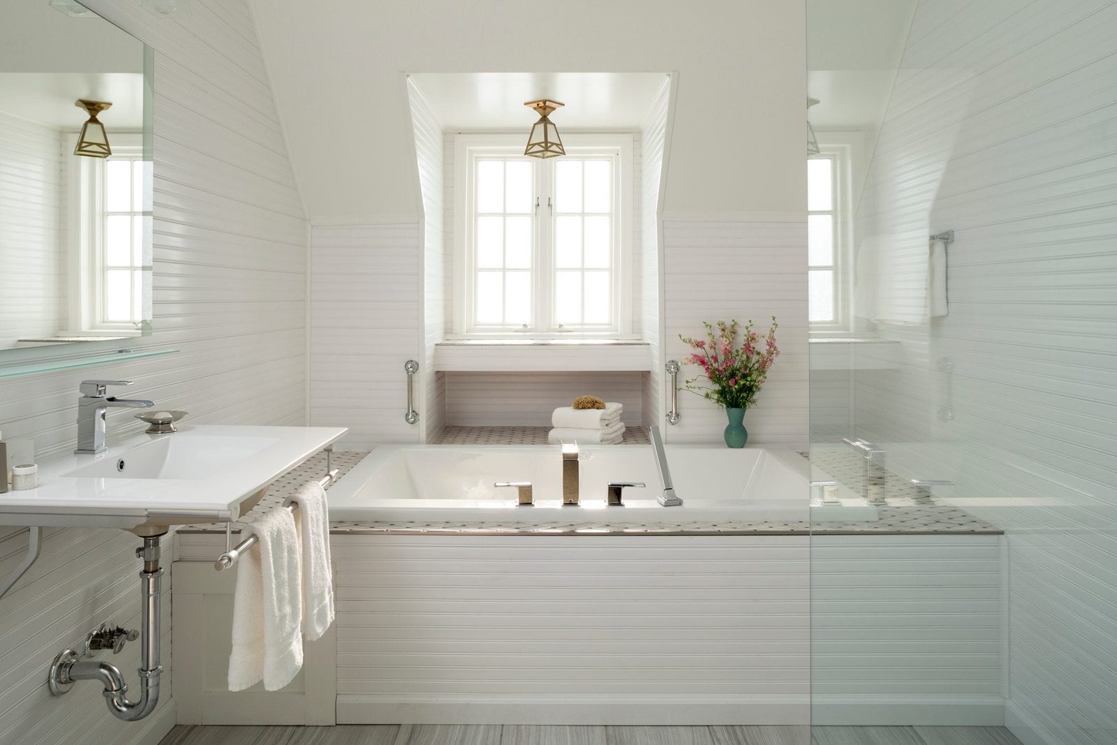 A white bathroom features a bathtub and sink, with towels hanging nearby, surrounded by light tiles and a window displaying flowers outside.