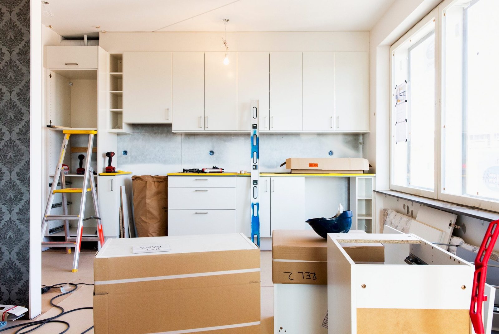 A ladder stands beside white cabinets being installed in a bright kitchen, with cardboard boxes and tools scattered across the unfinished fl
