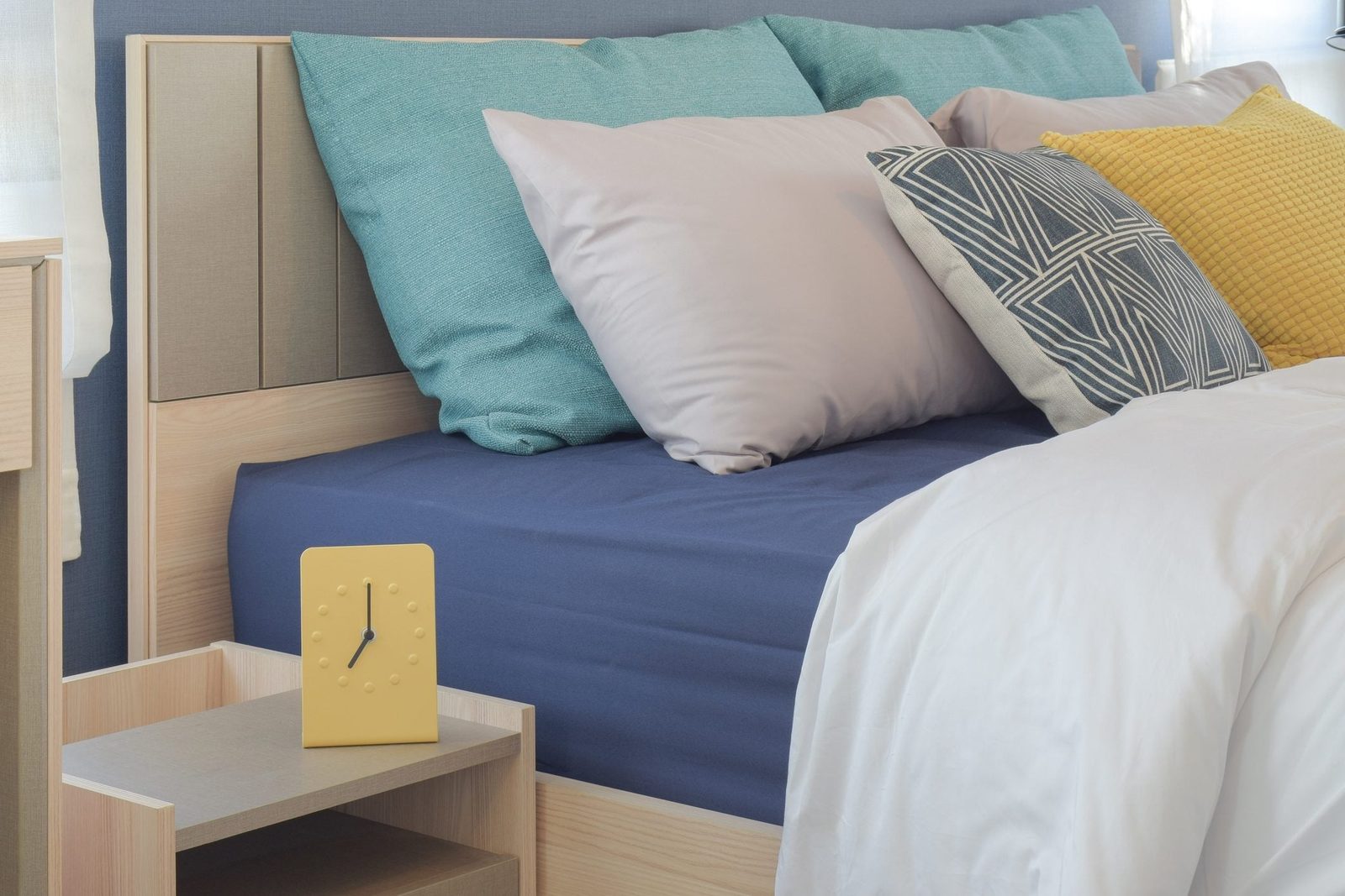 A bed with various pillows arranged on it, alongside a small yellow clock on a wooden nightstand in a neatly organized bedroom.