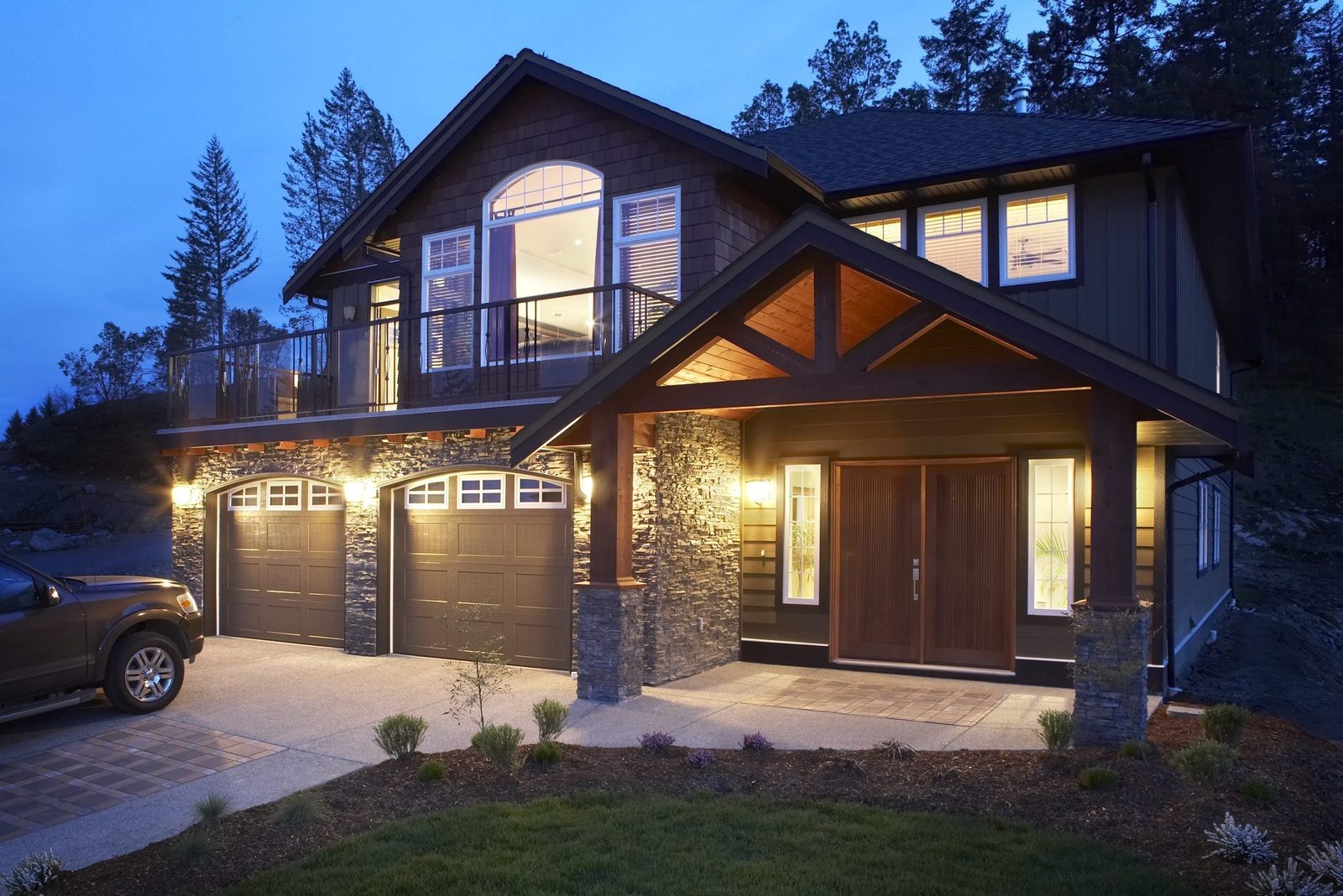A modern two-story house shines at dusk, featuring a dark exterior, large windows, a stone-accented garage, and landscaping with small plant