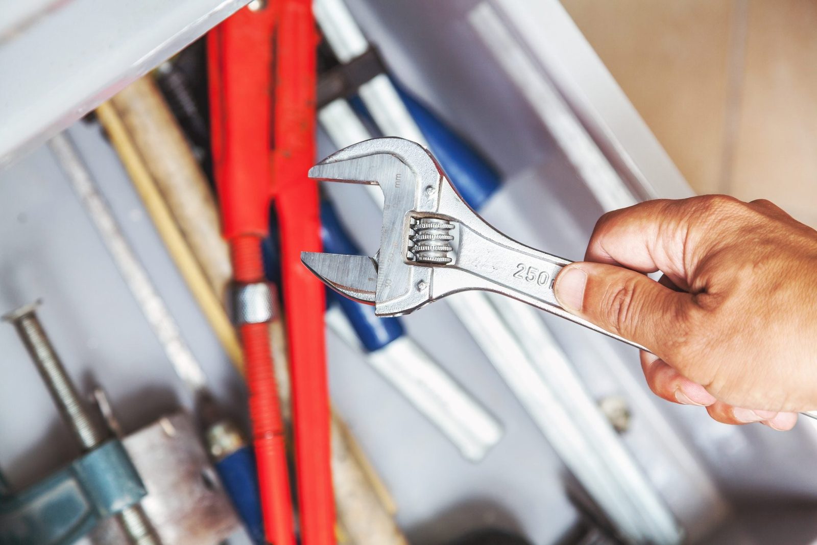 A hand grips a metallic adjustable wrench above a cluttered toolbox filled with various tools, including red pliers and metallic rods.