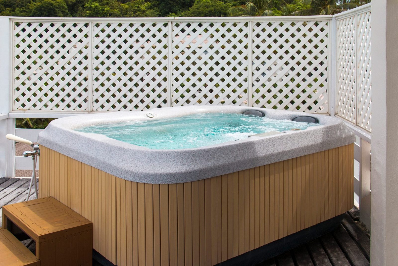 A hot tub bubbles, inviting relaxation, positioned on a wooden deck surrounded by a white lattice fence, with green trees visible in the bac