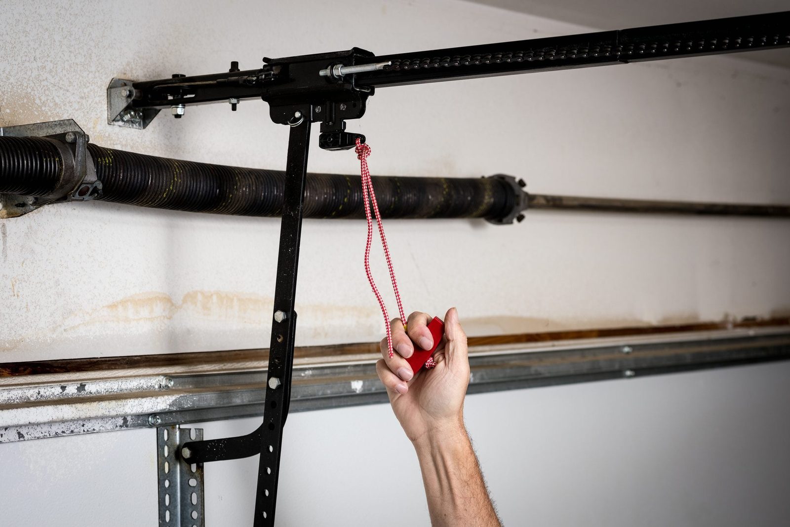 A hand pulls a red rope connected to a garage door mechanism, situated above a dark pipe, against a textured, white wall.