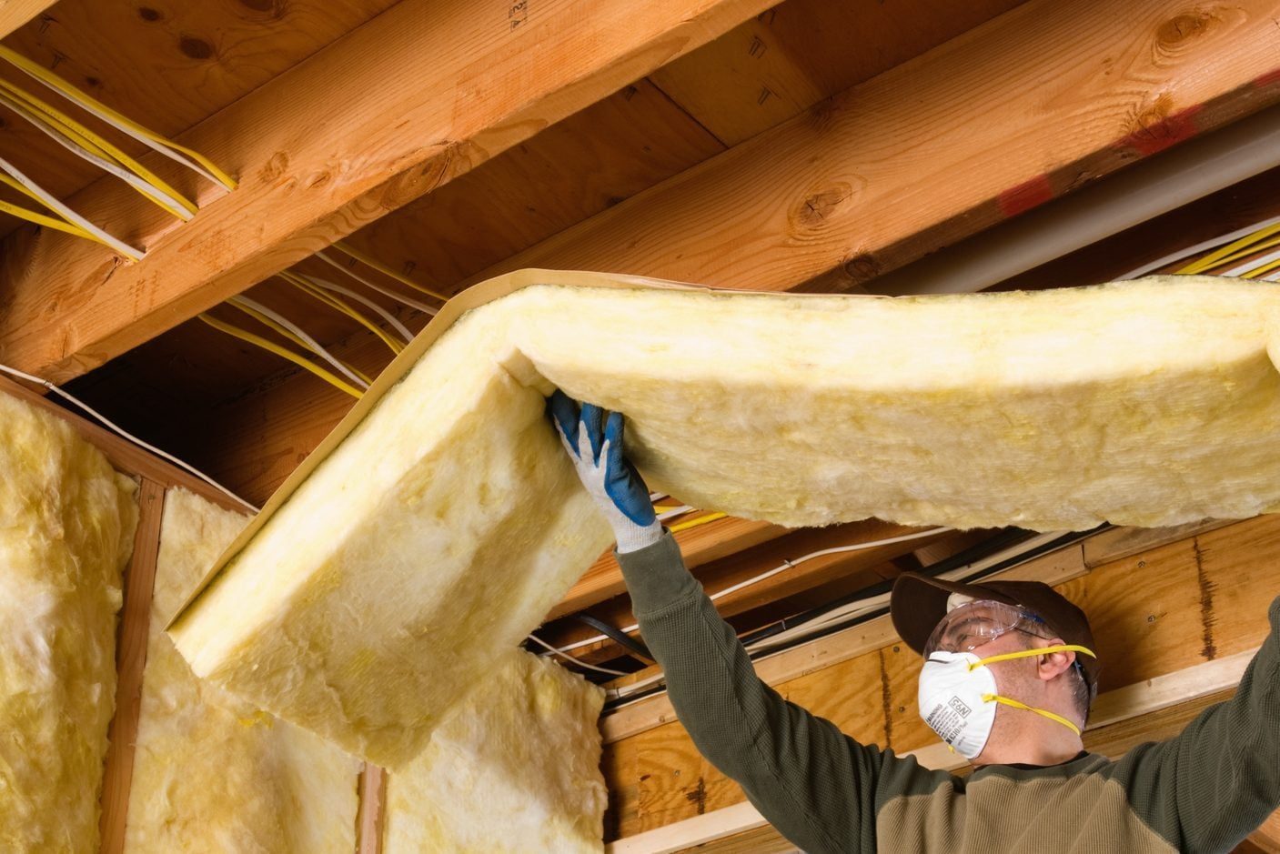 A worker lifts a large piece of insulation into a wooden ceiling, surrounded by exposed beams and electrical wiring.