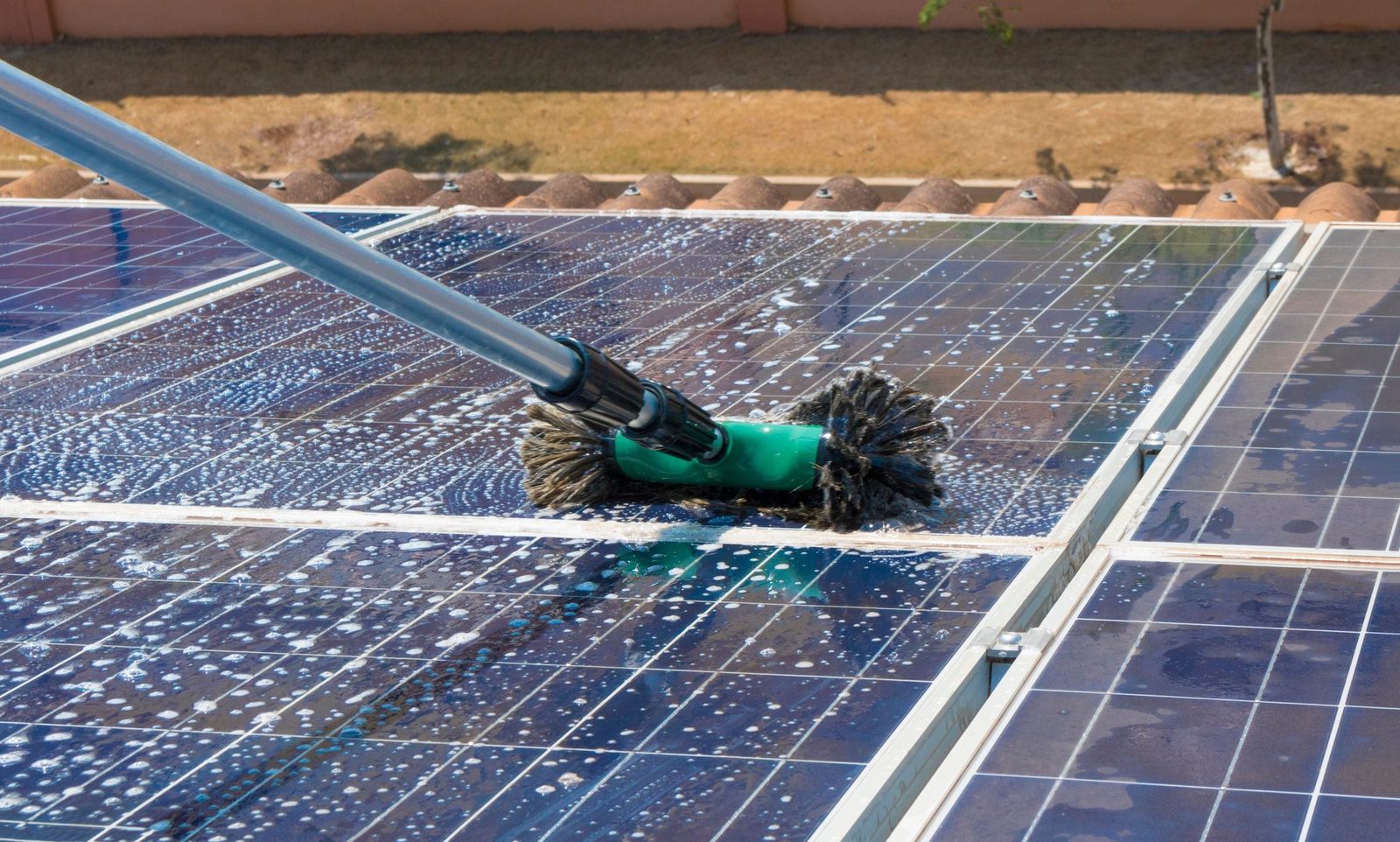 A cleaning brush scrubs a solar panel, generating foam, on a rooftop surrounded by dry grass and a wall, under a clear sky.