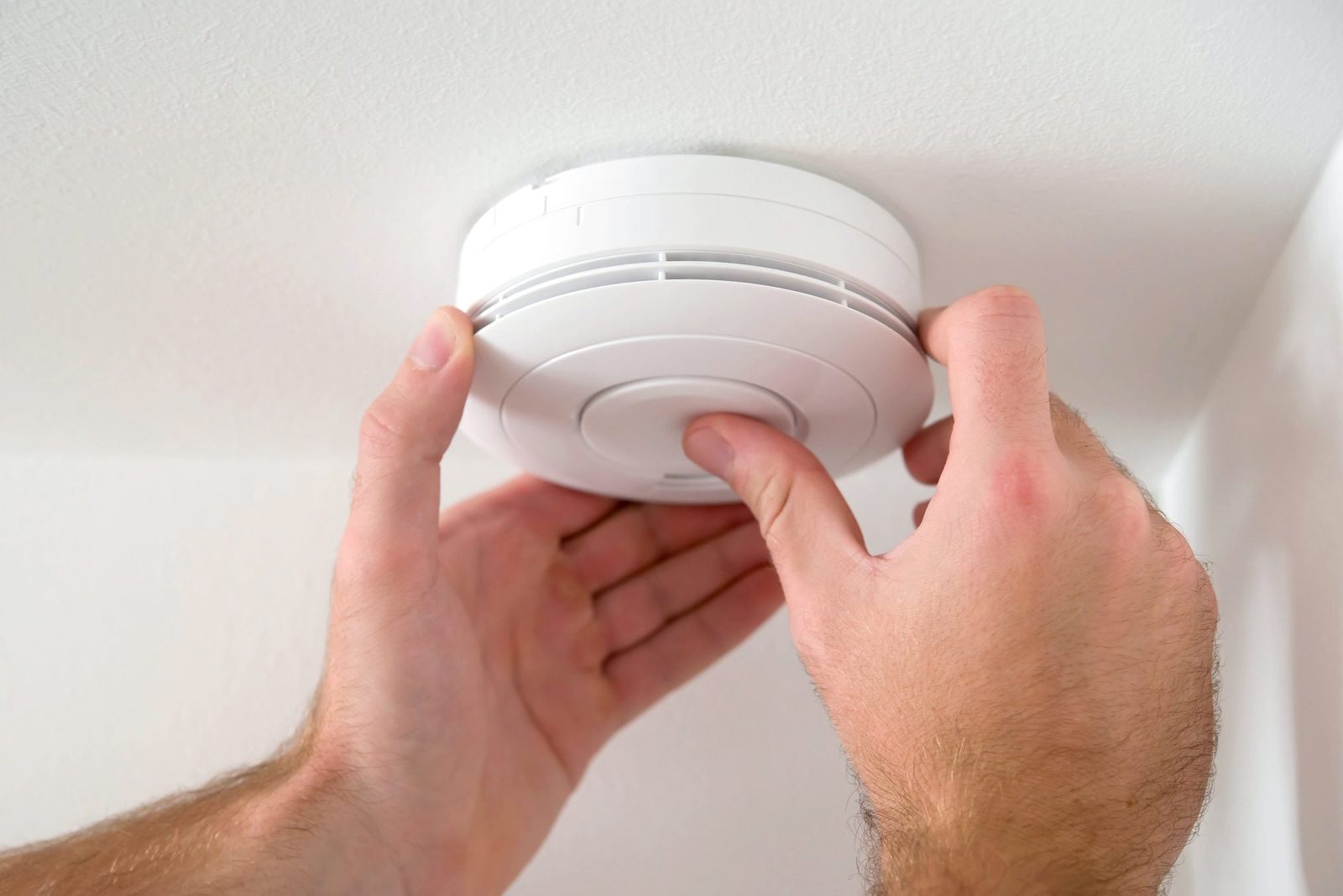 A person is installing a smoke detector on a white ceiling, ensuring it's securely fitted in a residential environment.