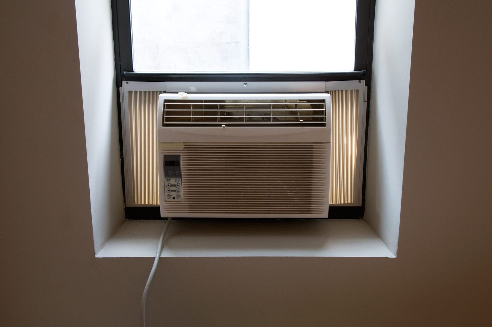 An air conditioning unit is installed in a window, emitting cool air into a light-colored, minimalistic room with plain walls.