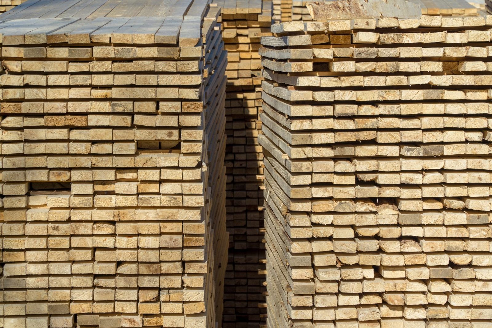 Stacked wooden planks are neatly arranged, creating parallel rows with an opening in between, situated in a bright, outdoor environment.
