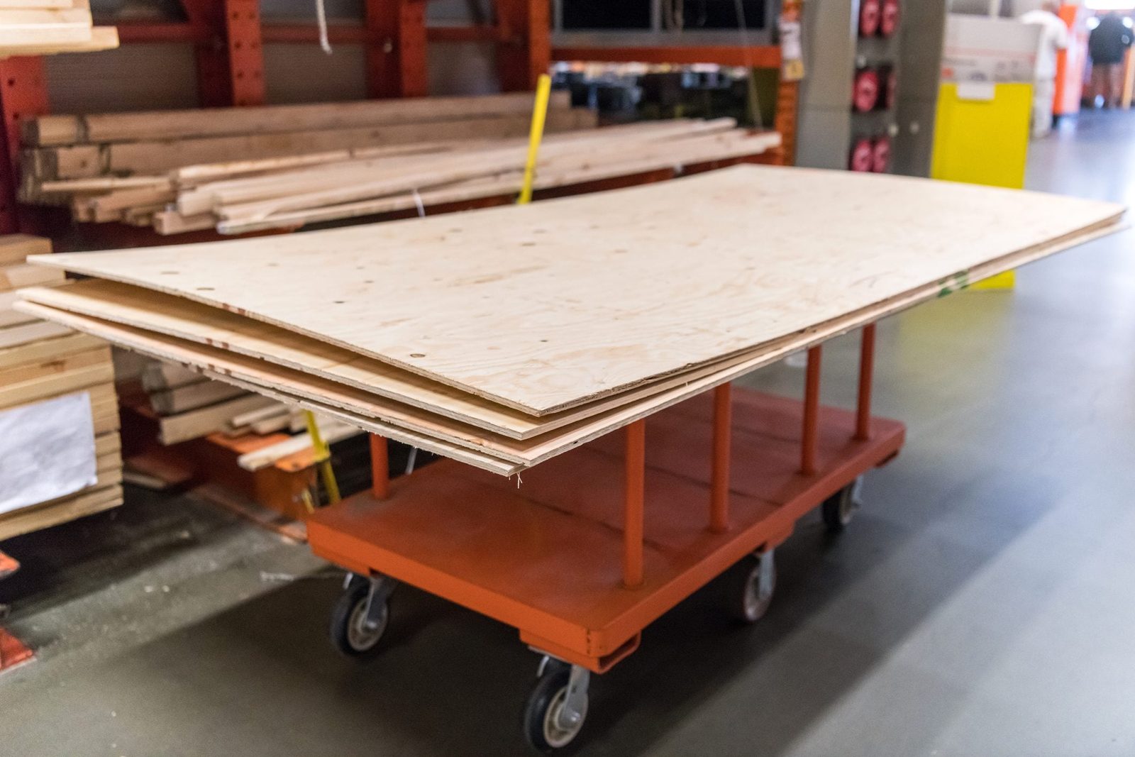 A cart carries stacked plywood sheets in a home improvement store aisle, surrounded by wooden planks and hardware, on a smooth concrete floo