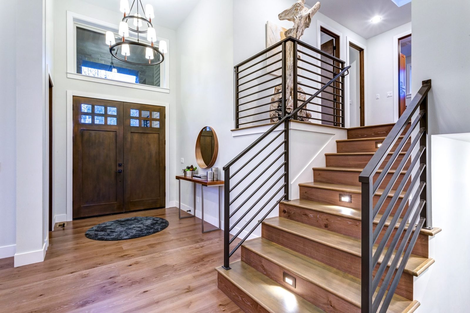 A staircase ascends beside a wall, illuminated by lights, leading to rooms while a wooden front door stands at the entrance, framed by a mod