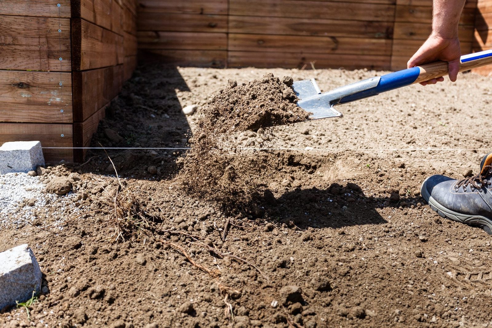 A person using a shovel lifts soil, scattering it onto the ground, near wooden borders within a dry, sunny outdoor area.