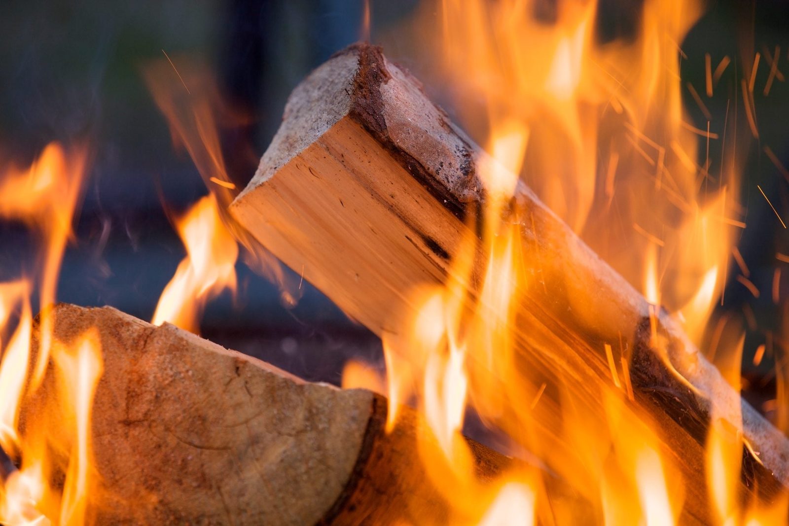 A log burns, surrounded by bright flames and sparks, creating a warm and dynamic atmosphere in a natural setting.