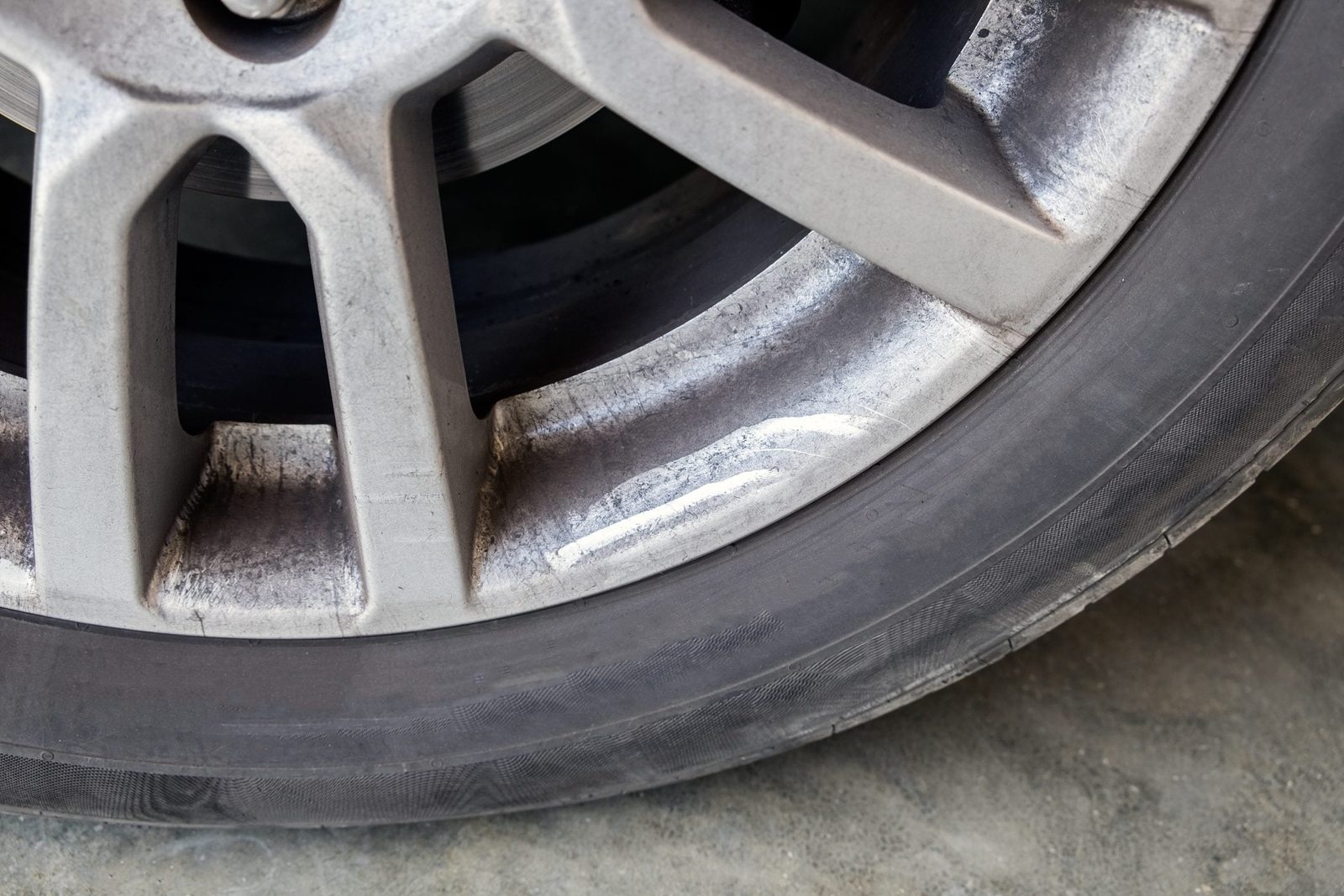 A dirty car wheel leans sideways, showing scratched metal and a worn tire against a concrete surface, suggesting it may require maintenance.