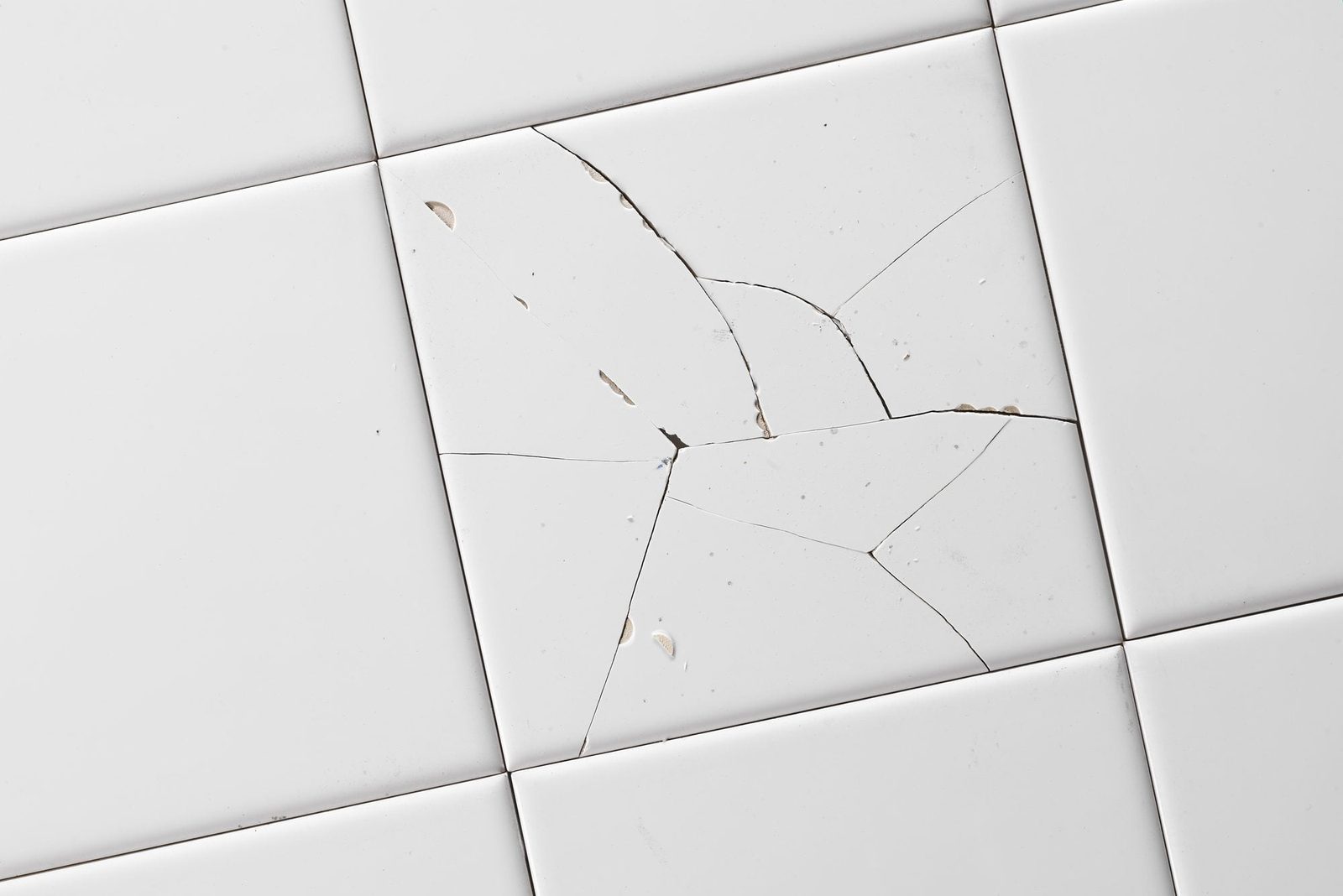 A cracked white tile lies on a floor, with visible fractures and blemishes, suggesting damage in a clean, light environment.