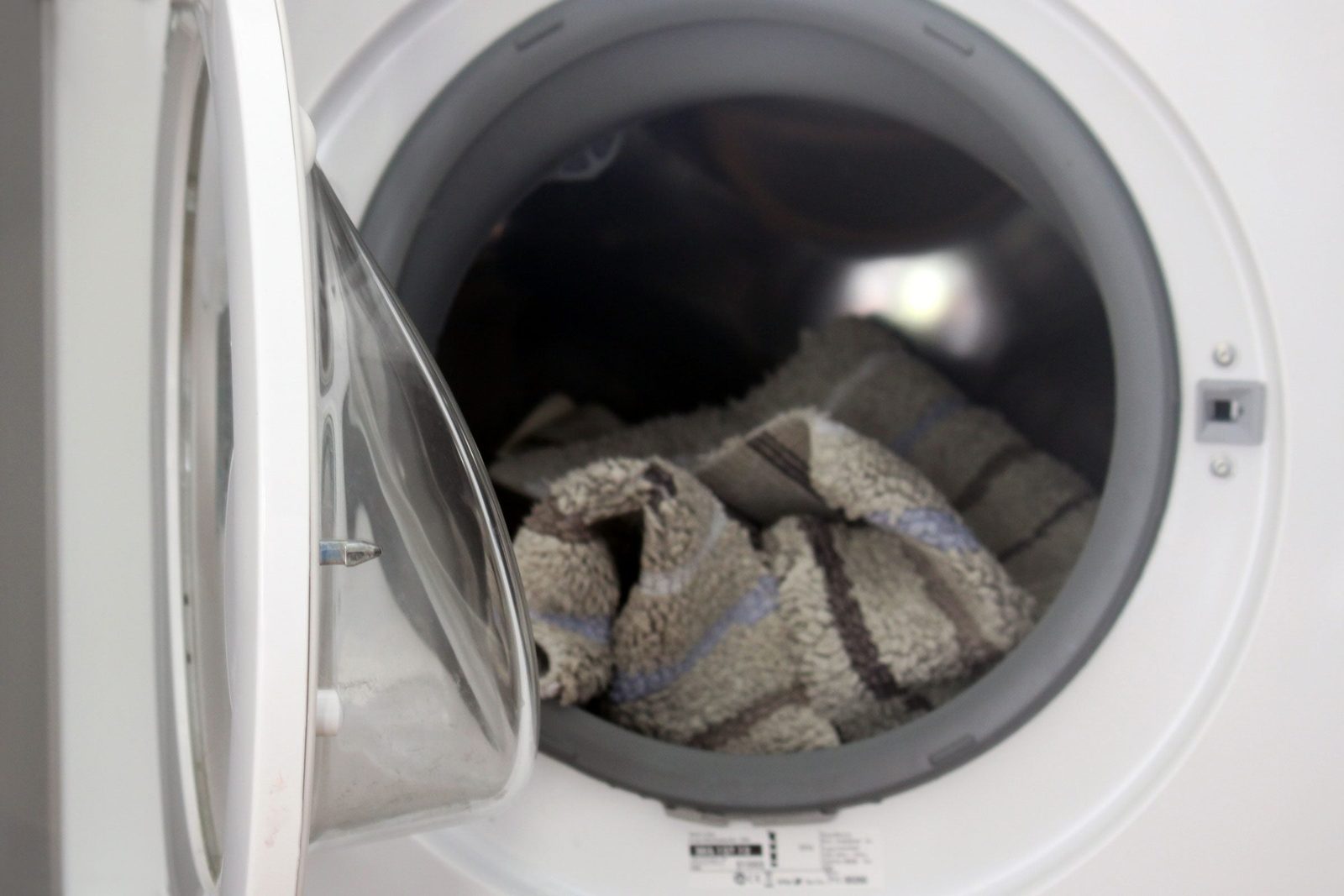 A washing machine door is open, revealing a patterned towel inside, resting against the drum in a bright, minimal laundry space.