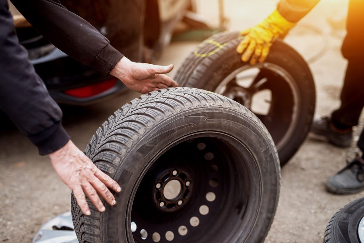 Two hands lift a car tire, while other tires and a vehicle are nearby on a work surface, indicating a tire change or maintenance activity.