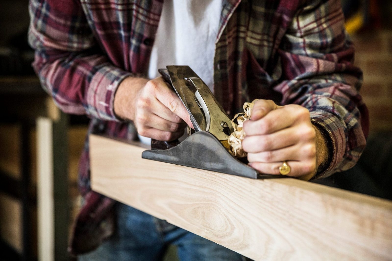 A person uses a hand plane to smooth a wooden board, producing shavings in a workshop setting with tools visible in the background.