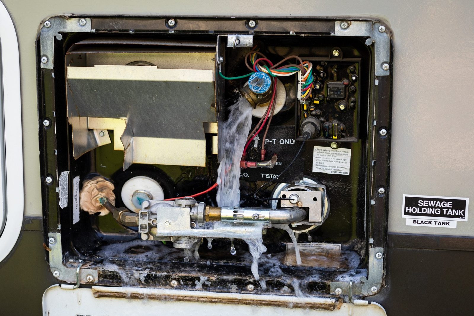 A sewage holding tank's access panel is open, water is flowing out through a connected valve, surrounded by wires and a black tank label.