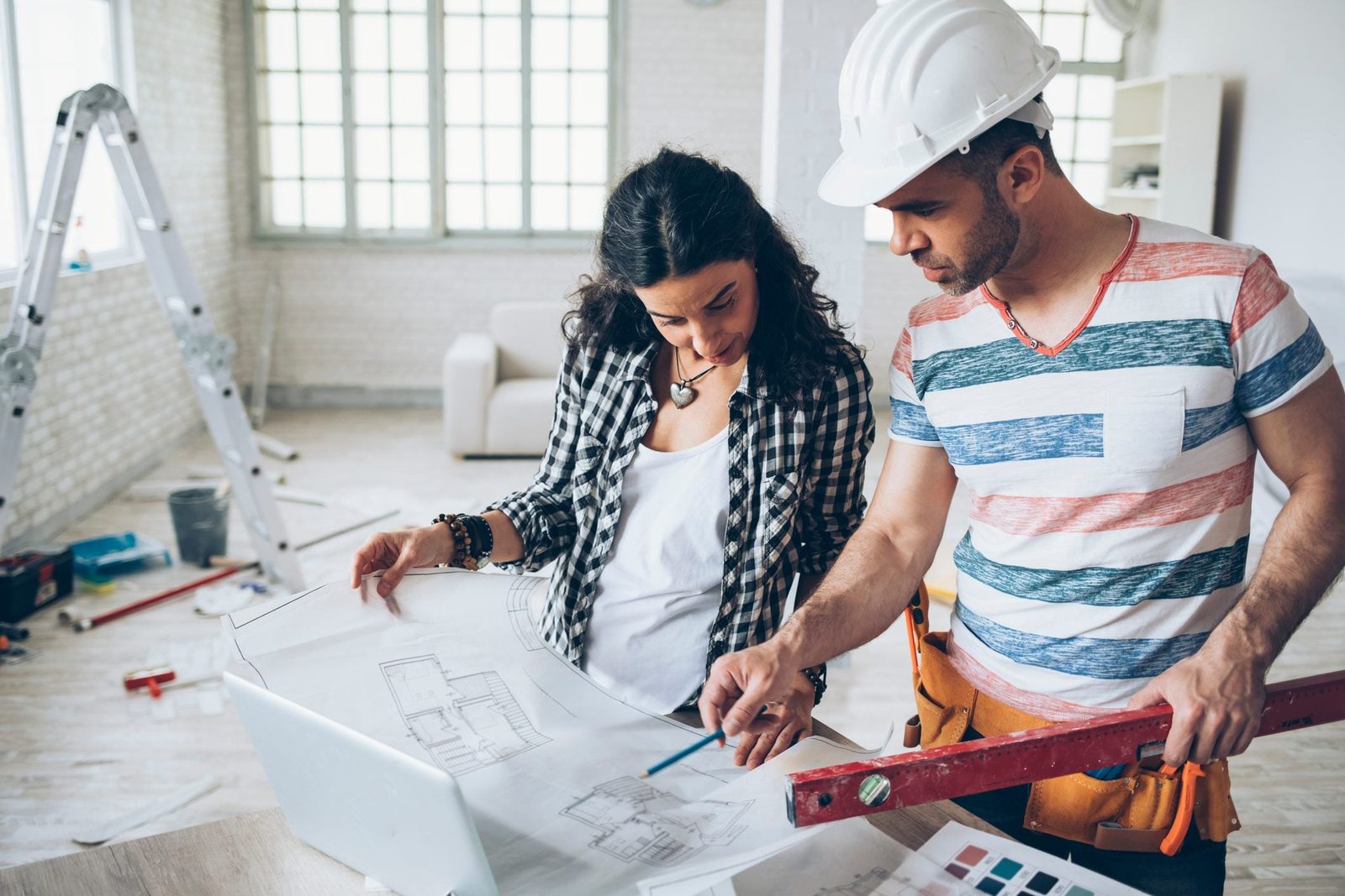 Two individuals examine blueprints on a table, discussing construction plans amid a partially furnished, light-filled room with tools and a