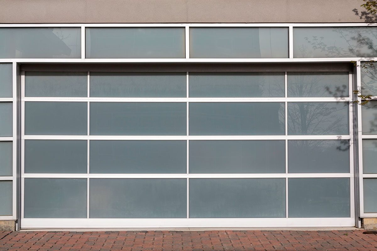 A garage door with large, frosted glass panels is closed, set within a modern building's gray wall with a brick pathway in front.
