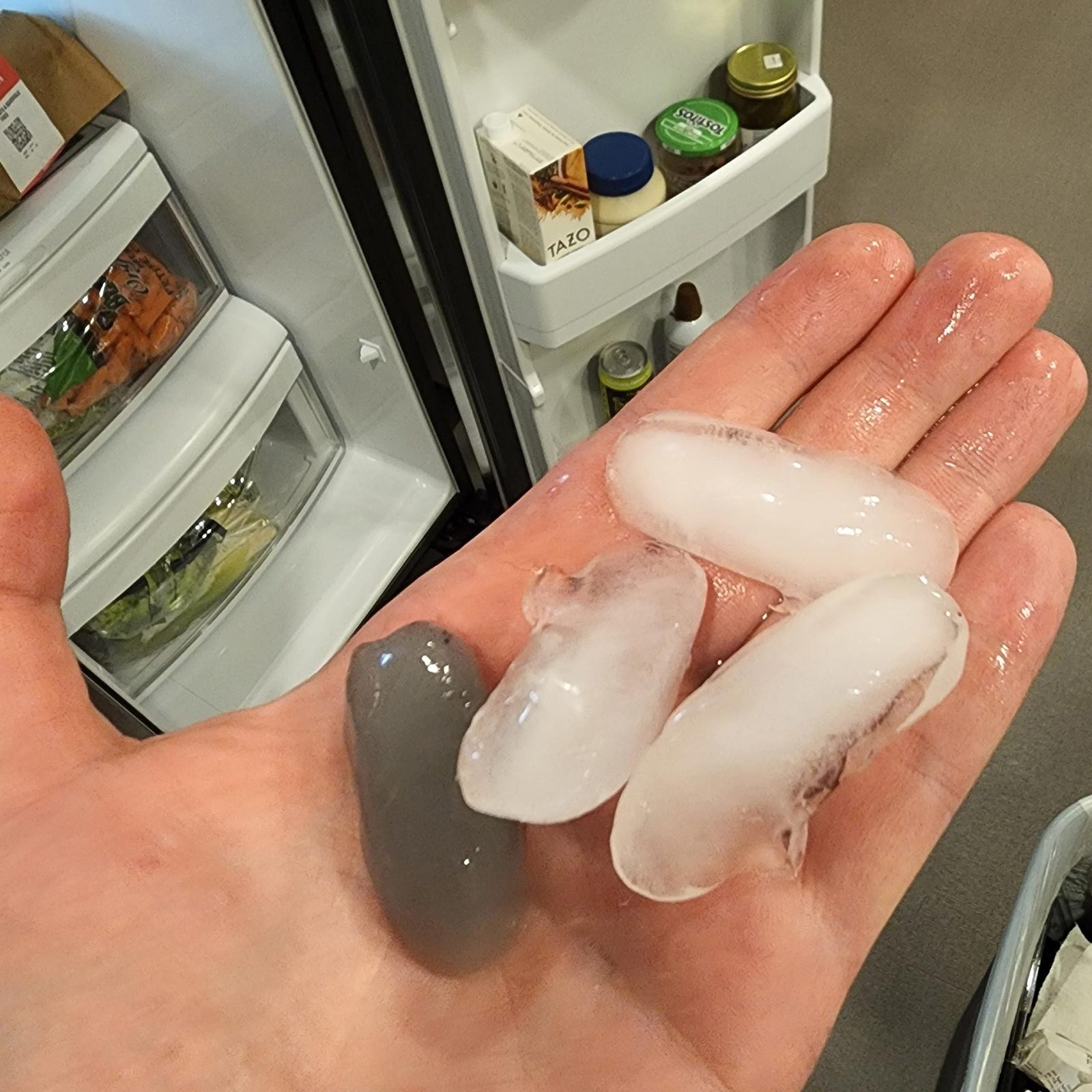 A hand holds four ice cubes of varying sizes and colors, while an open refrigerator background contains food items and drinks.