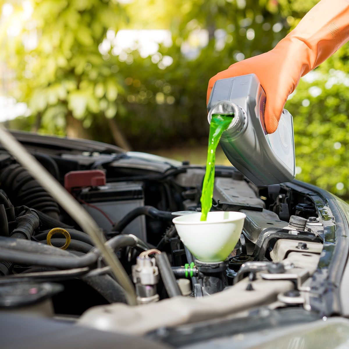 A person in orange gloves pours green fluid into a funnel above a car engine, surrounded by greenery and bright sunlight.