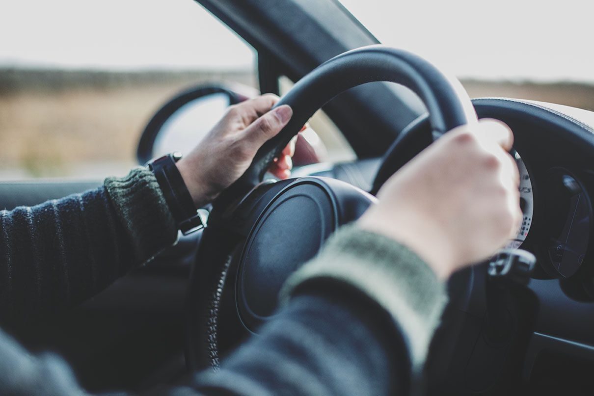 Hands grip a steering wheel, maneuvering the car along a road with a blurred landscape visible through the windows.