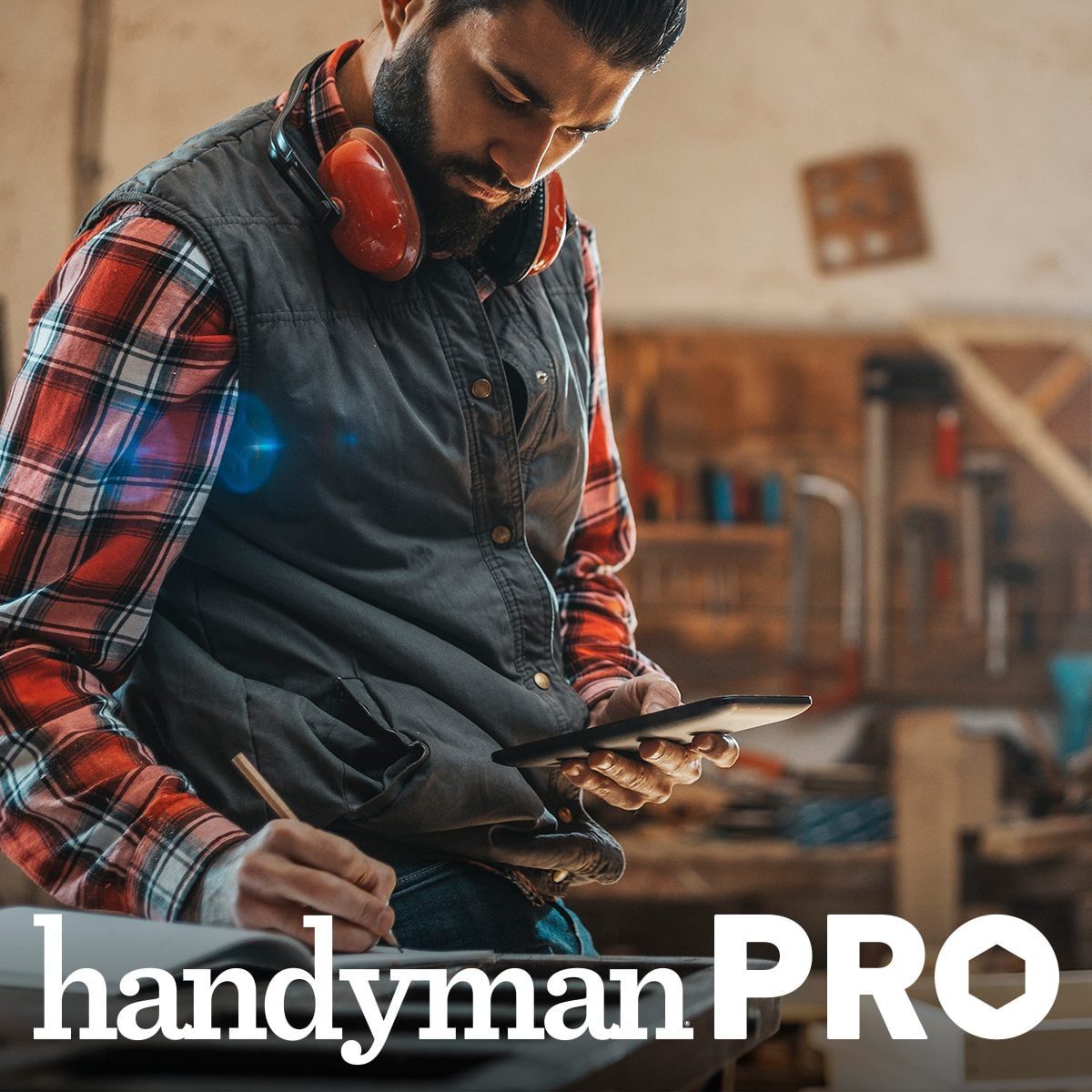 A man wearing safety headphones reviews plans on a tablet while seated at a workbench in a workshop filled with tools and woodwork materials