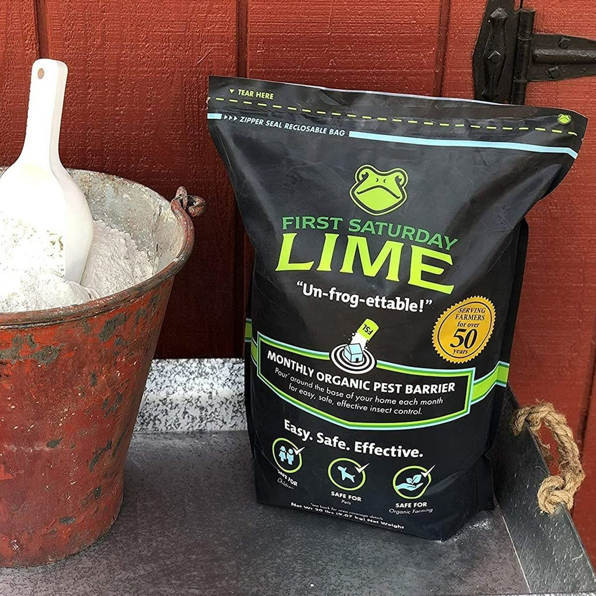 A black bag of First Saturday Lime stands beside a rusted bucket filled with white powder, outdoors against a red wooden backdrop.