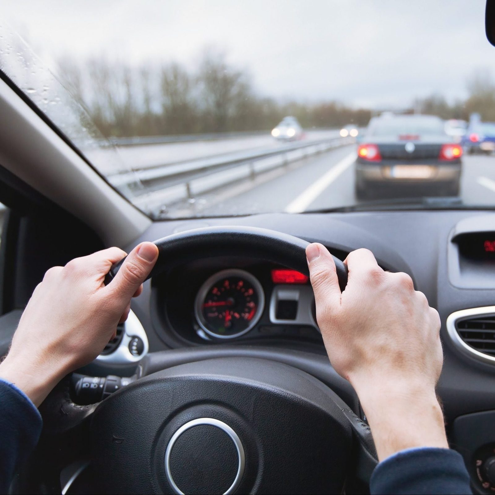 Hands grip a steering wheel while driving on a wet highway, with surrounding cars partially visible in overcast weather.
