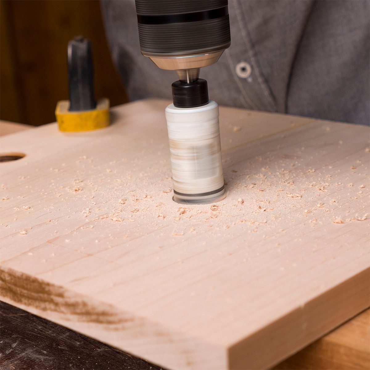 A drill is spinning above a wooden surface, creating shavings as it carves out a hole, with tools visible in the background.