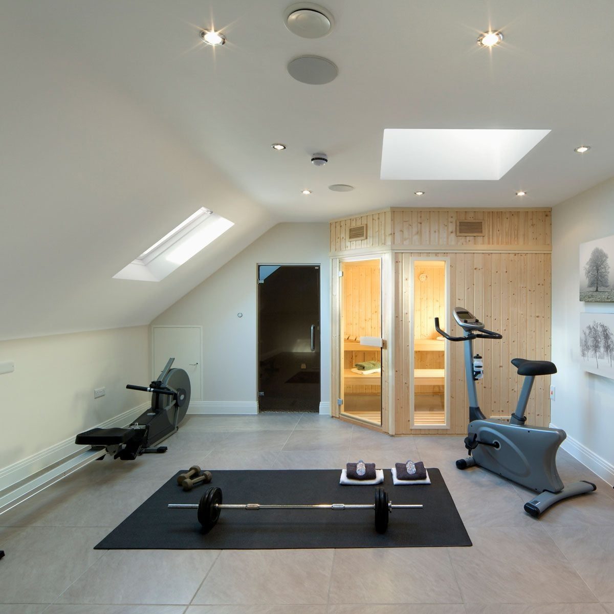 A home gym features a wooden sauna, exercise equipment, and a barbell on a mat, illuminated by recessed lighting under a sloped ceiling.
