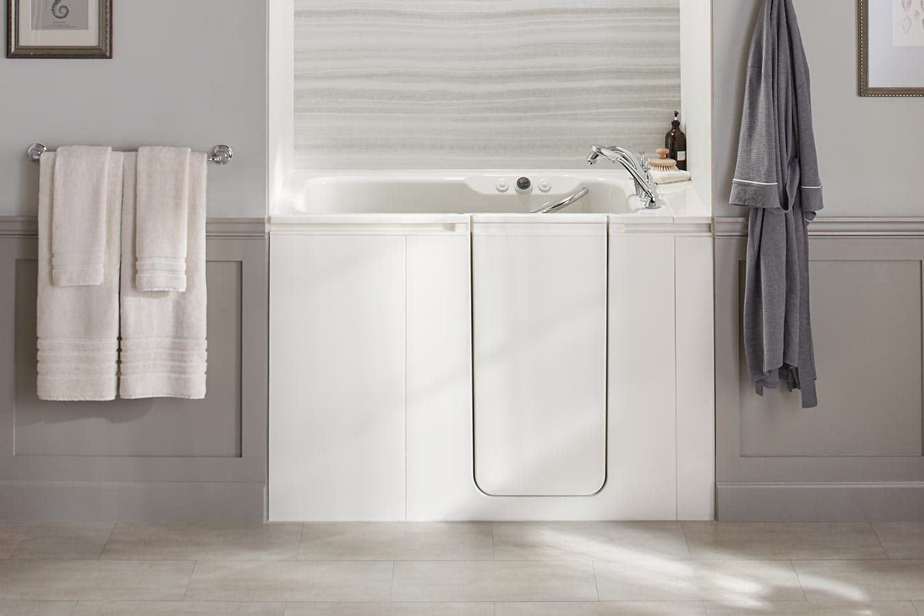 A white walk-in bathtub sits against a neutral wall, with towels hanging nearby and a bathrobe draped, suggesting a relaxing bathroom enviro