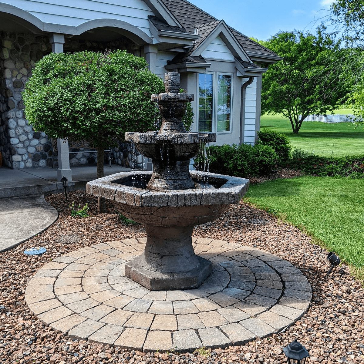 A stone fountain with three tiers flows water, surrounded by a circular paved area and landscaped greenery, next to a house on a sunny day.