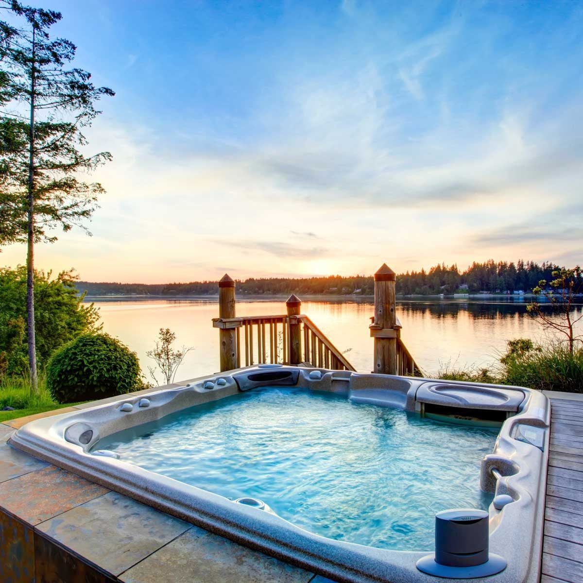 A hot tub filled with water sits on a deck, overlooking a calm lake at sunset, surrounded by trees and a wooden staircase leading down to th