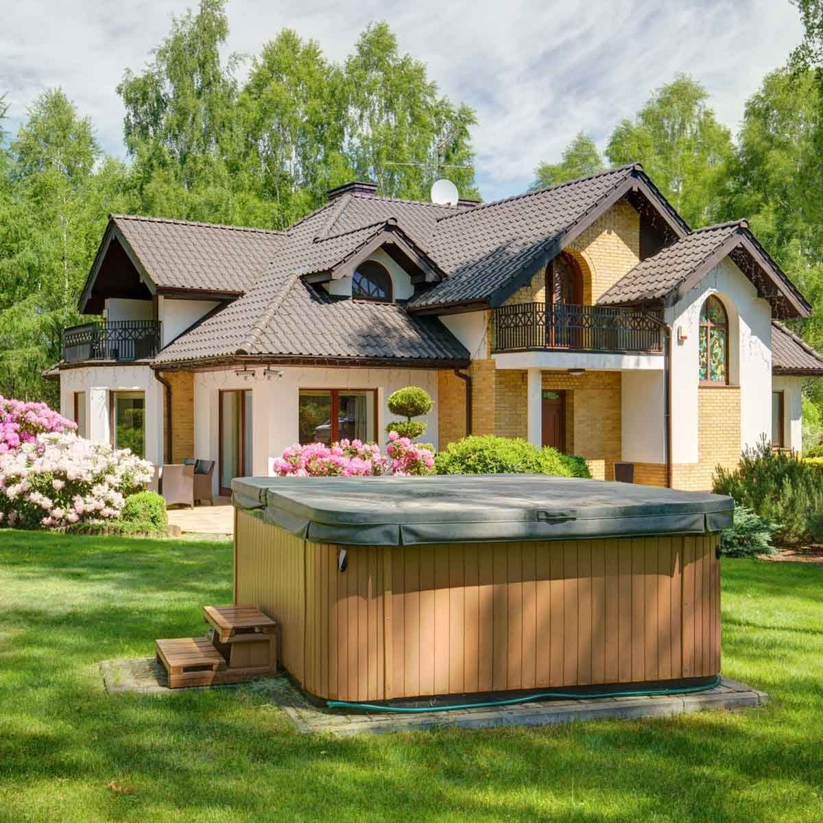 A wooden hot tub sits on grass, surrounded by flowering shrubs, with a two-story house featuring a sloped roof and large windows nearby.