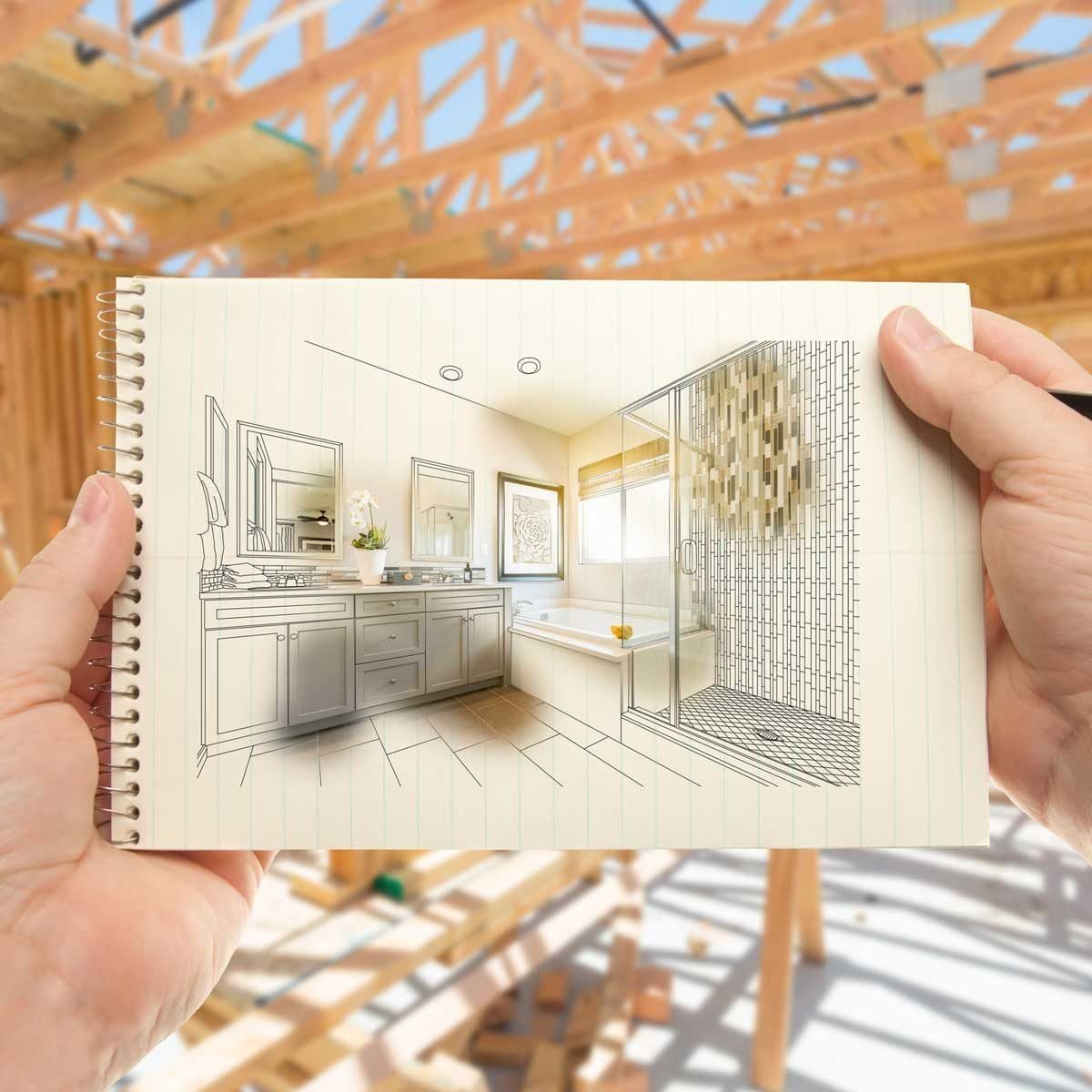 A hand holds an illustrated bathroom design on a notebook. The background shows a construction site with exposed wooden beams and unfinished