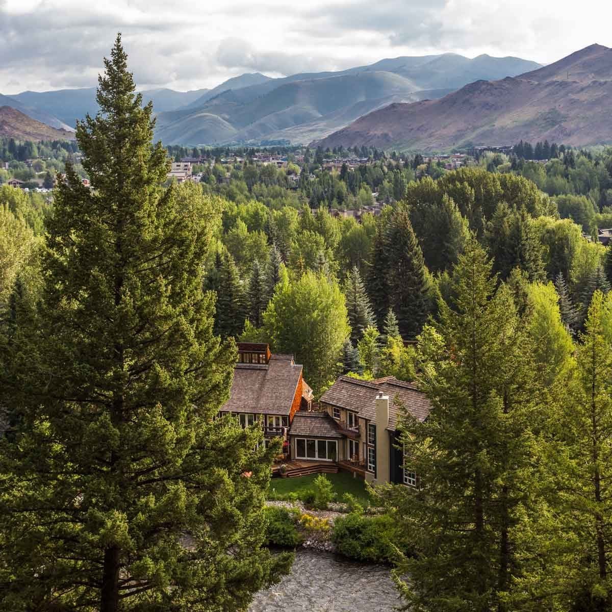A cluster of houses sits near a flowing river, surrounded by dense trees and mountains under a partly cloudy sky.