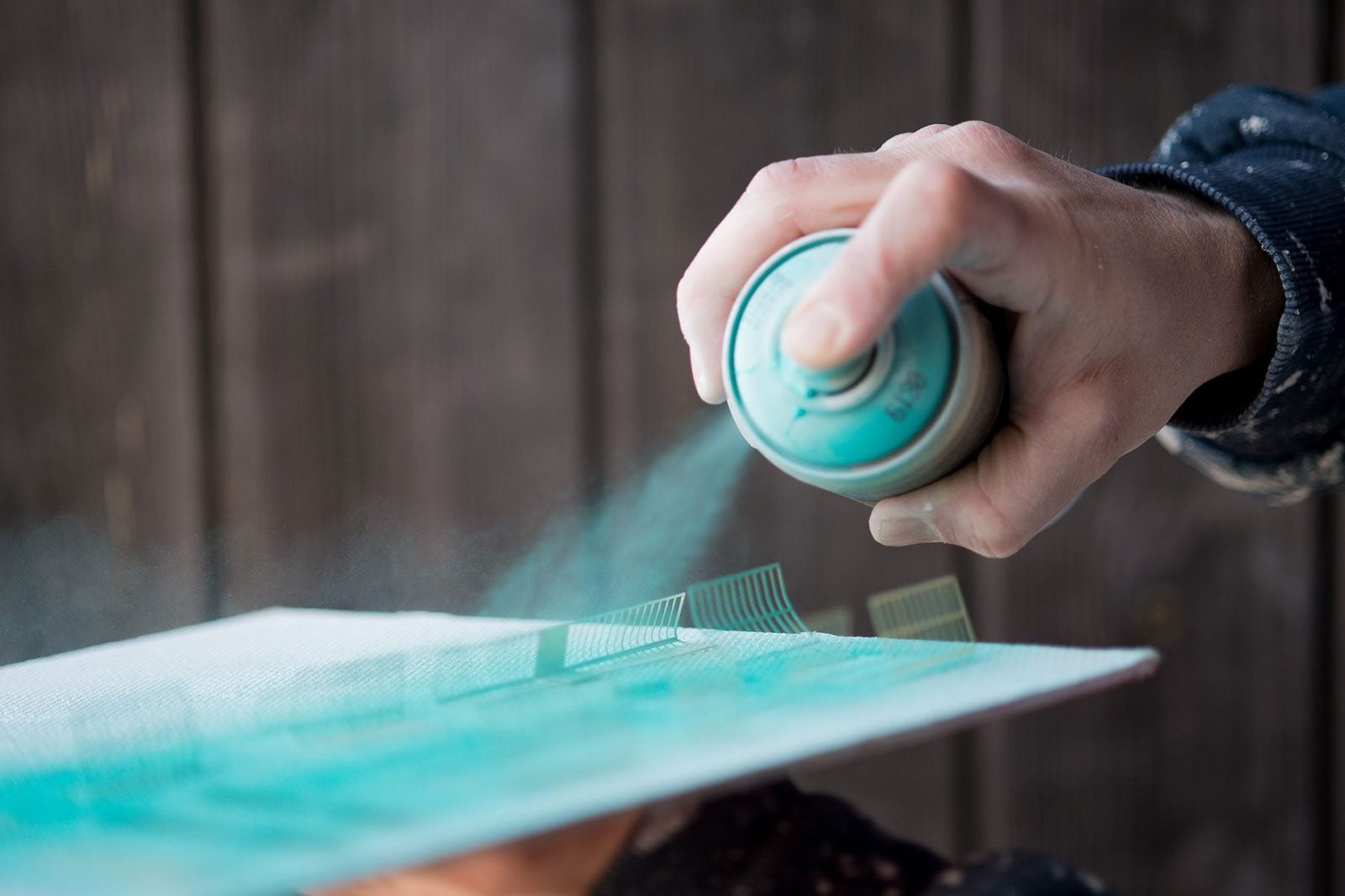 A hand sprays turquoise paint onto a textured canvas, creating a mist in front of a rustic, wooden backdrop.