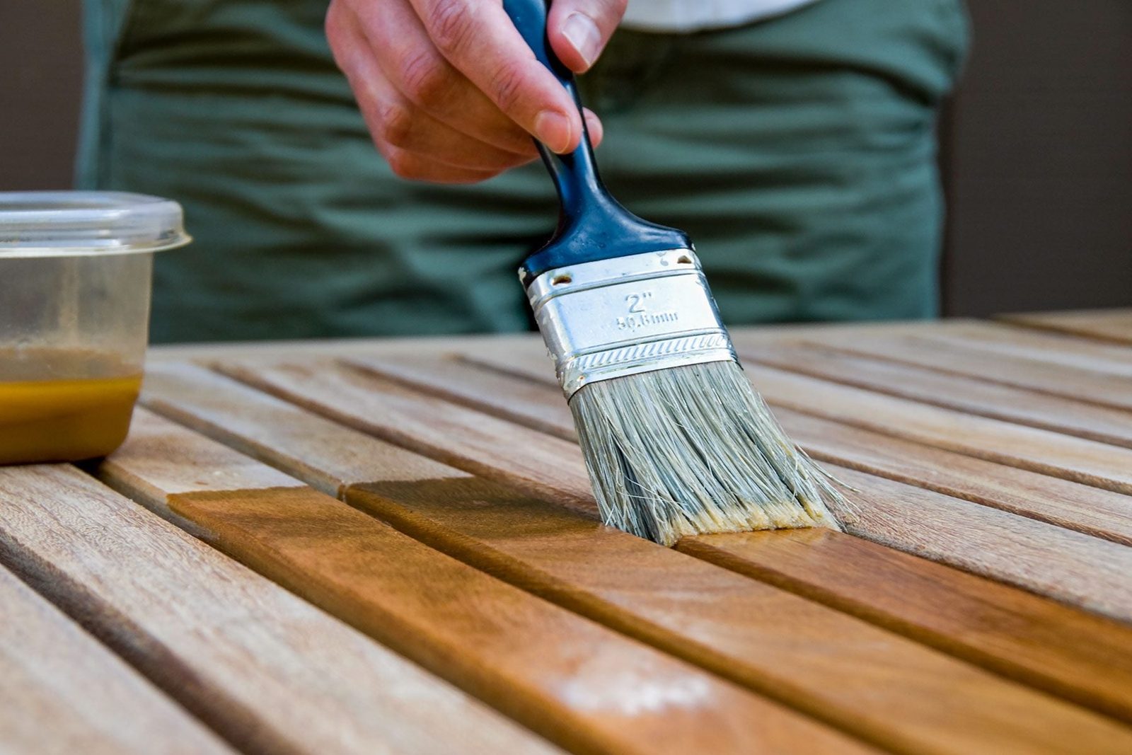 A paintbrush applies finish to wooden slats on a table, with a container of stain nearby, in a well-lit outdoor setting.
