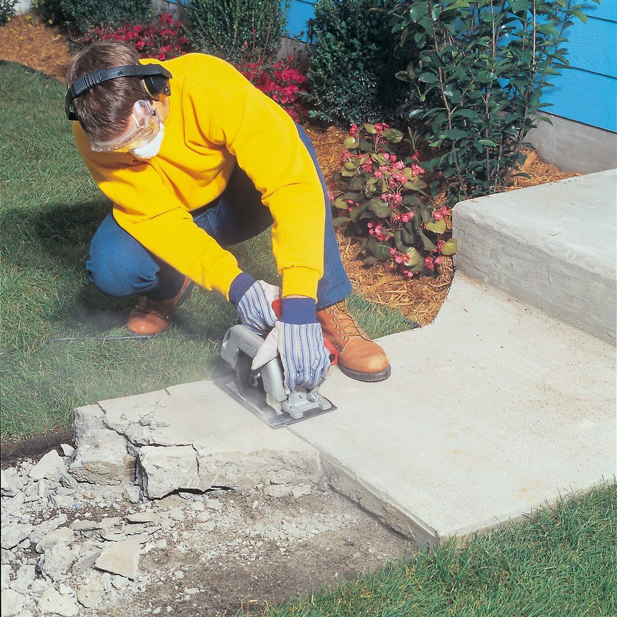 A person wearing protective glasses, ear protection, gloves, and a yellow jacket is using a circular saw to cut through a concrete slab next to a garden bed with green plants and