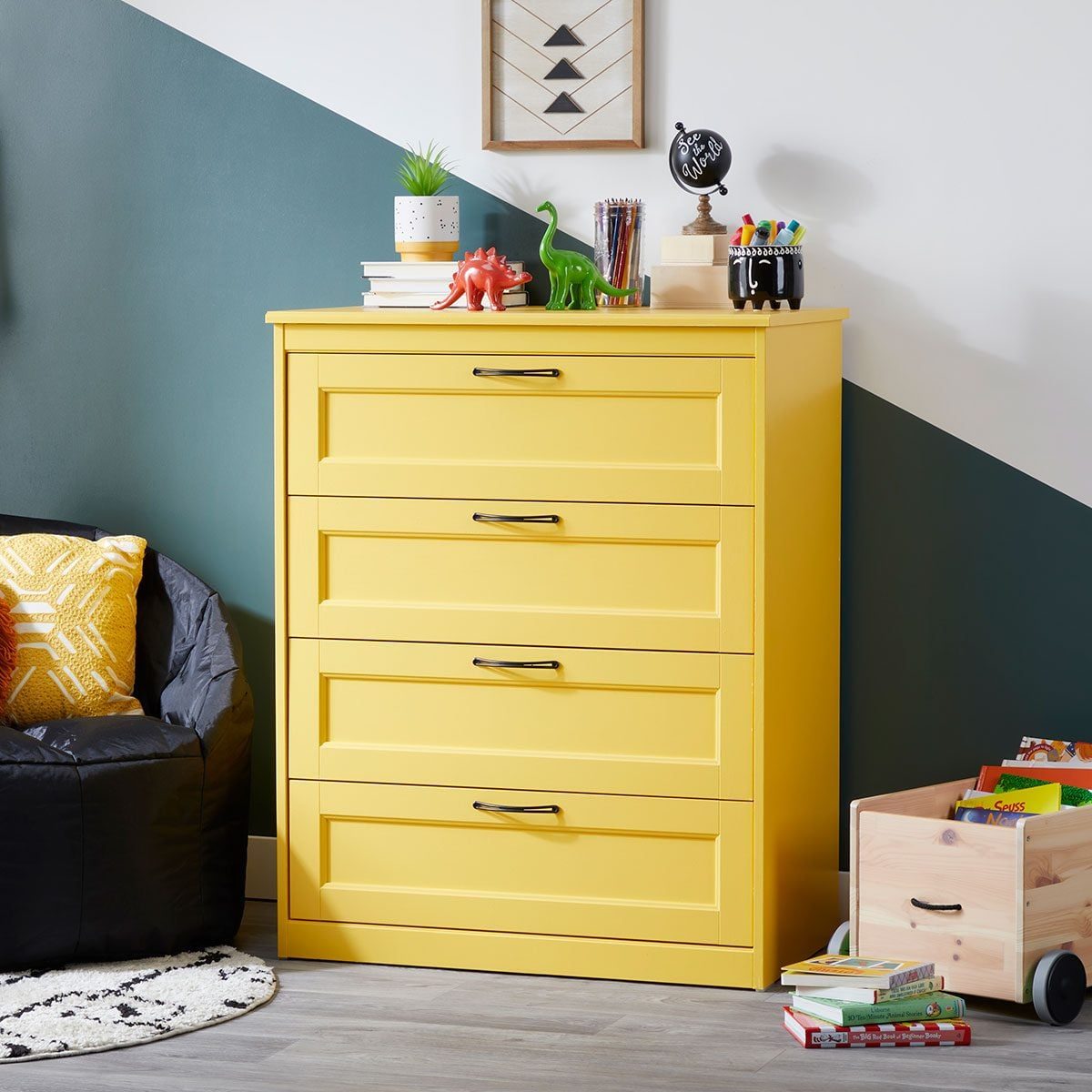 A yellow dresser stands against a wall, holding plants and toys. A black bean bag chair and a wooden cart with books complement the playful
