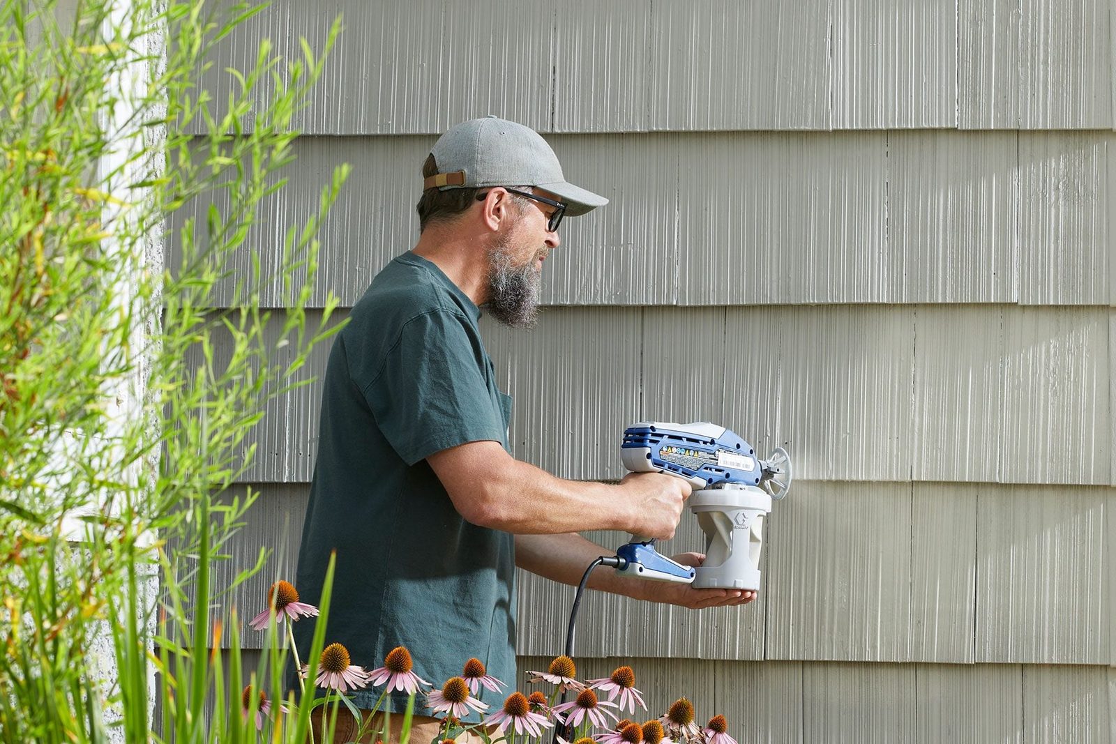 A man holds a spray painting tool, aiming it at a textured wall while surrounded by blooming flowers and greenery in a well-lit outdoor sett