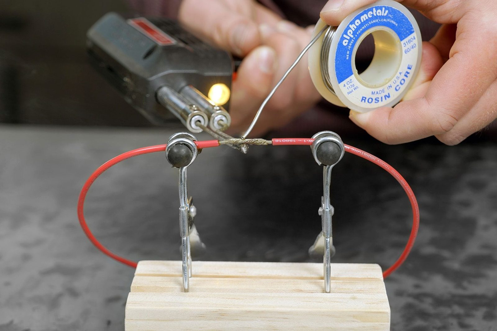 A soldering iron is being used to join two red wires, secured by clips on a wooden base, with solder being added from a spool.