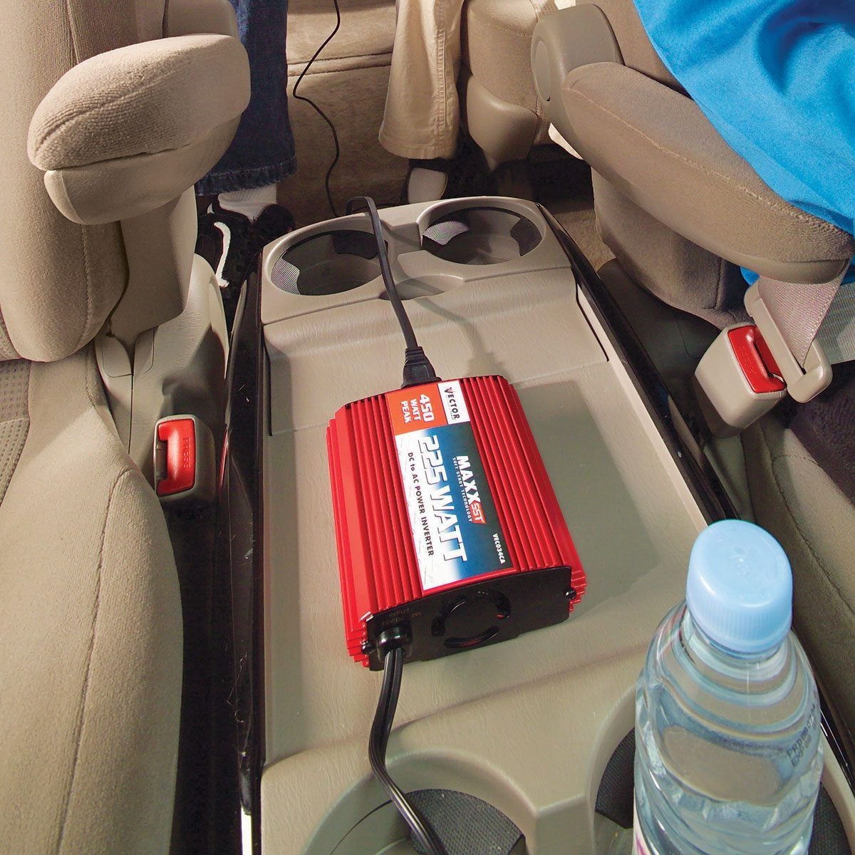 A red power inverter sits on a car's center console, connected to a power source, while a water bottle is nearby, indicating a travel contex