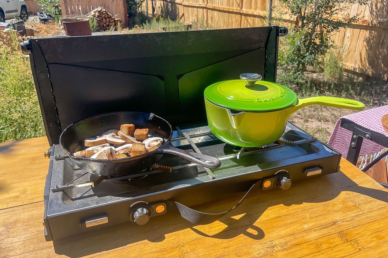 A portable camping stove on a wooden table holds a frying pan with mushrooms and tofu, and a green pot with a lid, outdoors near a wooden fence.